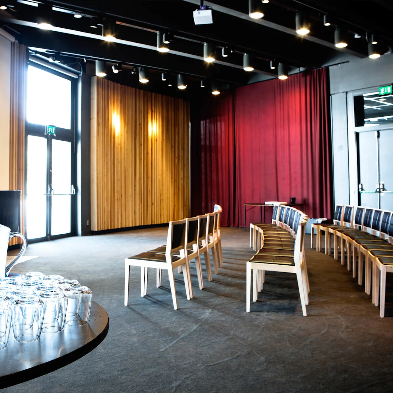 Pitchers of lemon water and glasses on a round table; in a conference room with wooden panels, chairs in rows facing a large screen, and red curtains. 