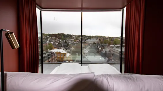 Seng med hvide puder vender mod et gulv-til-loft-vindue med udsigt over bylandskab med bygninger og grønne områder. Røde gardiner omgiver udsigten, og en messinglampe står ved siden af sengen.