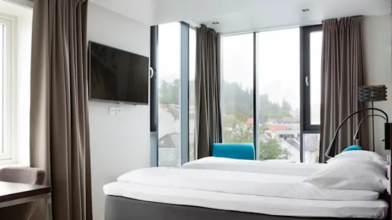 En seng med hvitt sengetøy er plassert mot store vinduer på hotellrommet, med utsikt over en by med trær og bygninger. En veggmontert TV og skrivebord er i nærheten.