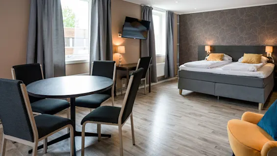 A modern hotel room features a double bed with yellow pillows, a round table with four chairs, a wall-mounted TV, and two large windows with gray curtains.