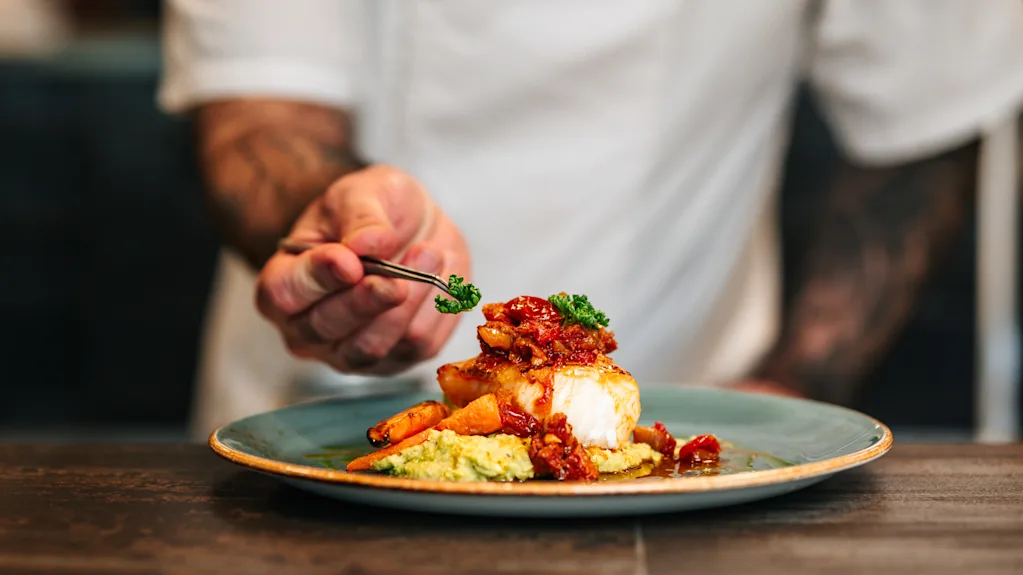 A chef using tweezers to carefully garnish a beautifully plated fish dish.