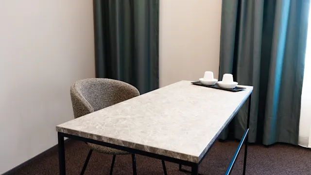 A rectangular marble-topped table positioned against a wall, with a single upholstered chair beside it. A tray holding two white cups sits on the table. Dark curtains hang in the background.