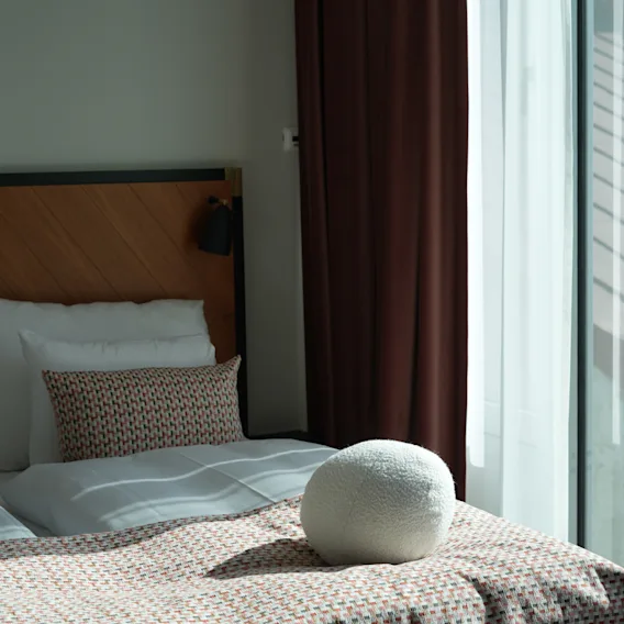 Hyggelig hotelseng med mønstret sengetøj og en rund hvid pude ved et lyst vindu