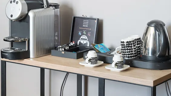 A coffee station with a Nespresso machine, capsules, kettle, and patterned cups sits on a wooden table. A framed poster reads: "Reload with L'OR Espresso Capsules" and info on espresso options.