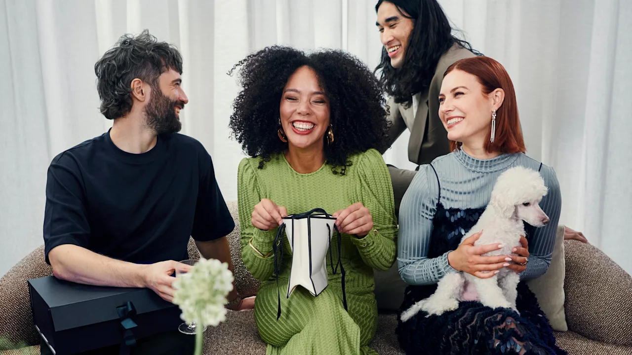 A group of friends gathered on a couch, one happily opening a gift while others smile and a small dog sits nearby.