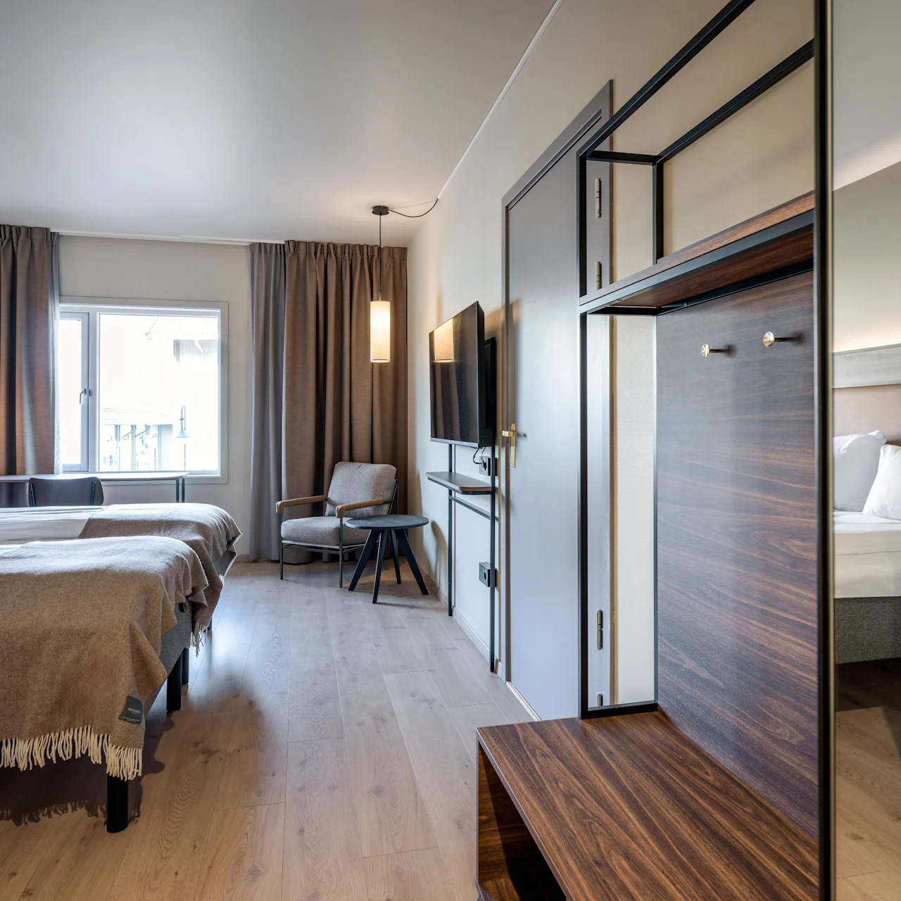 Two neatly made beds stand beside each other; a cozy chair and table are near a window. A flat-screen TV is mounted on the wall, enhancing the room's modern ambiance.