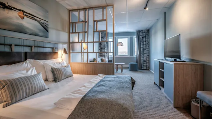 A bed with white linens and patterned pillows stands against a partition. A TV is on a cabinet opposite, with a sitting area near large windows.