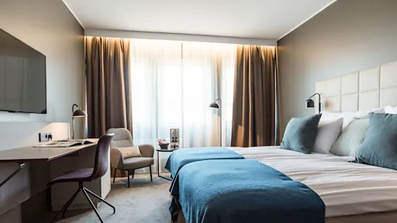 Bed sits neatly with blue blanket and pillows, beside a lamp-lit chair; desk with chair and book faces a wall-mounted TV, in a softly lit room.
