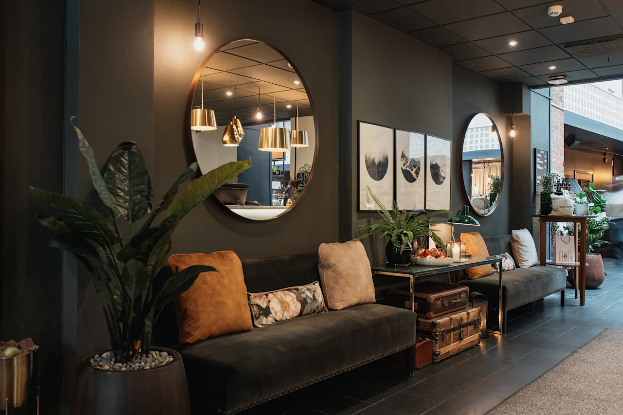Et hyggeligt og stilfuldt hotelloungeområde med mørke vægge, komfortable sofaer, dekorative planter, spejle og indrammet kunst.