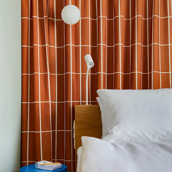 A cozy bedroom scene with a bed, a blue stool, and a patterned orange curtain. A reading lamp is visible.