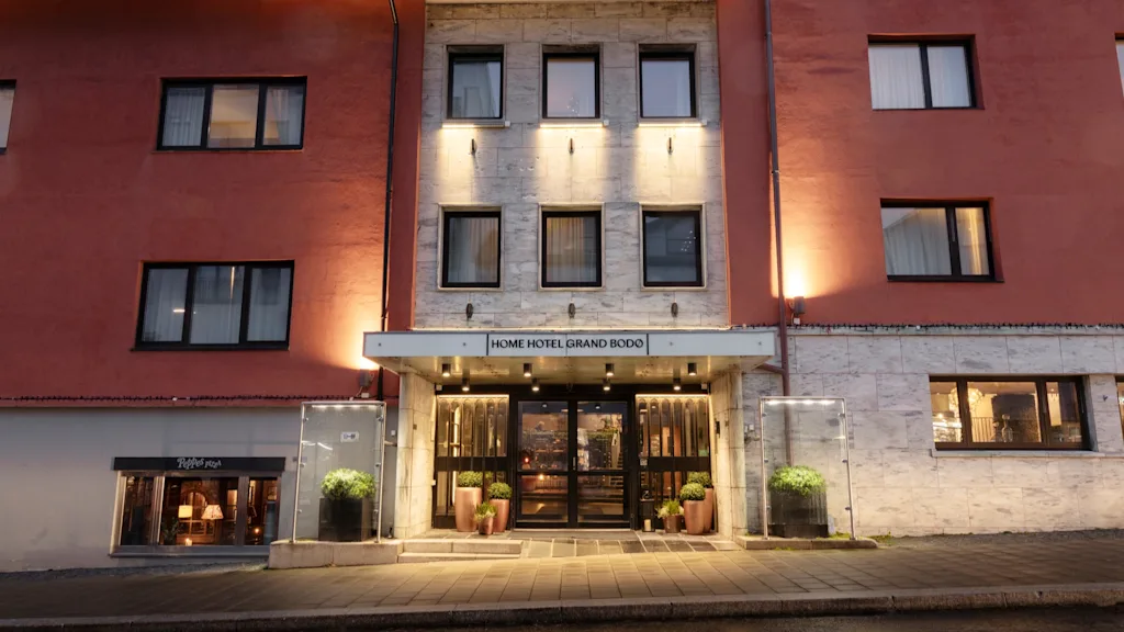 The illuminated entrance of a hotel building at dusk, with the sign HOME HOTEL GRAND BODO above the doors.