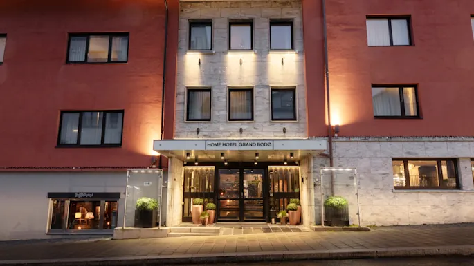 The illuminated entrance of a hotel building at dusk, with the sign HOME HOTEL GRAND BODO above the doors.