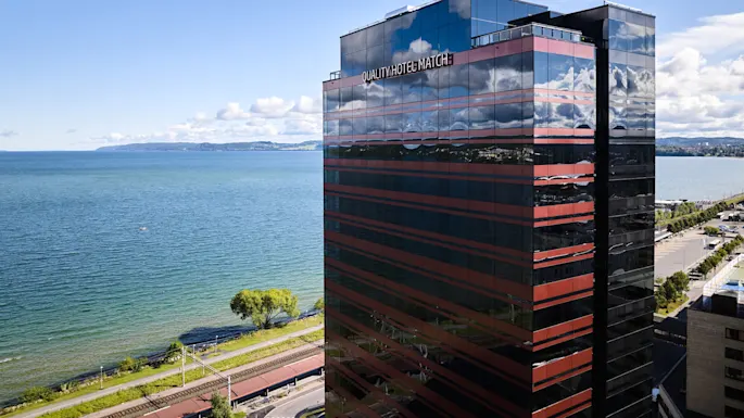 En høy bygning merket "QUALITY HOTEL MATCH" står ved siden av en stor, rolig innsjø under en klar blå himmel, med fjerne fjell og en tre-kantet strand i bakgrunnen.