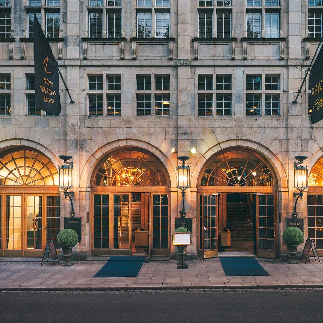 A grand building facade features three arched wooden entrances, illuminated by amber lights. Two flags hang vertically, reading “HOTEL CRISTINA TEATRO” and “TEATRO”. Mannequins display clothes in the left shop window.