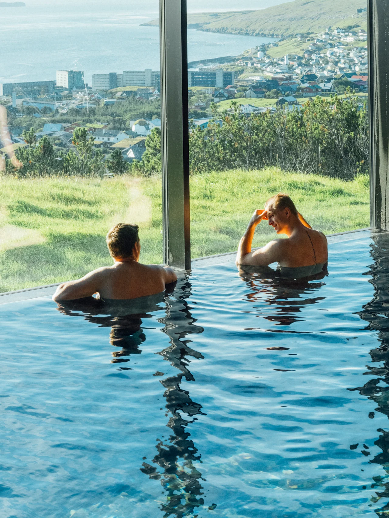 Two people relaxing in an infinity pool with a panoramic view of a coastal town and green landscape.
