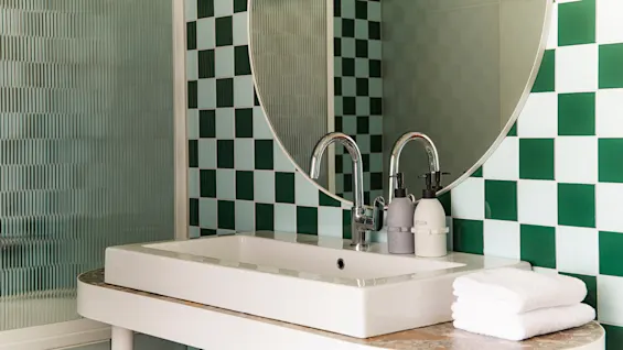 En vask med to blanke haner står under et rundt spejl. Flasker mærket "SOAP" og "LOTION" står på bordet med grønne kvadratiske vægfliser omkring.