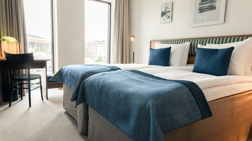 Et lyst hotellrom med to senger, blå detaljer, et skrivebord ved vinduet og kunst på veggen, for et komfortabelt opphold.