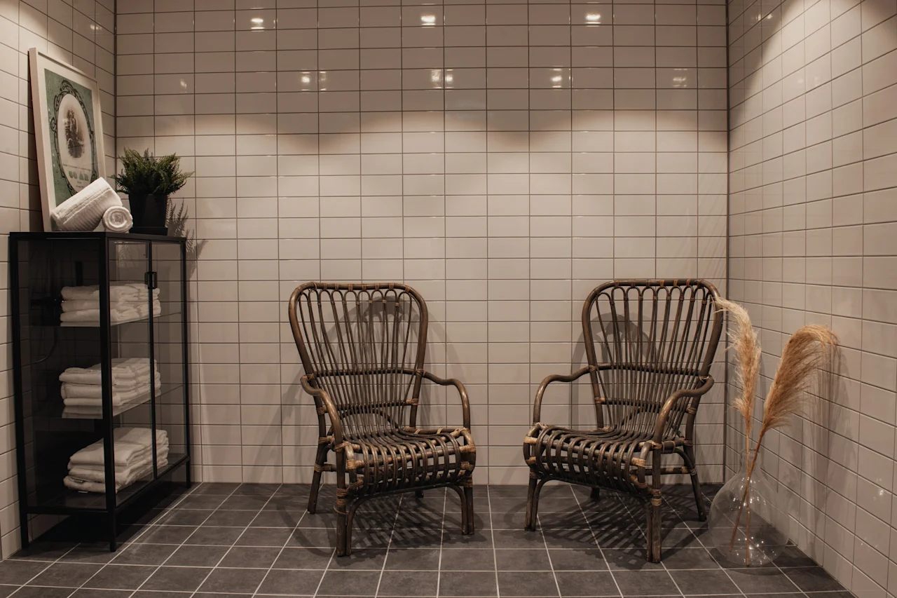Et roligt rum med flisebelagte vægge og gulv, med to fletstole, et skab med håndklæder og dekorative planter.
