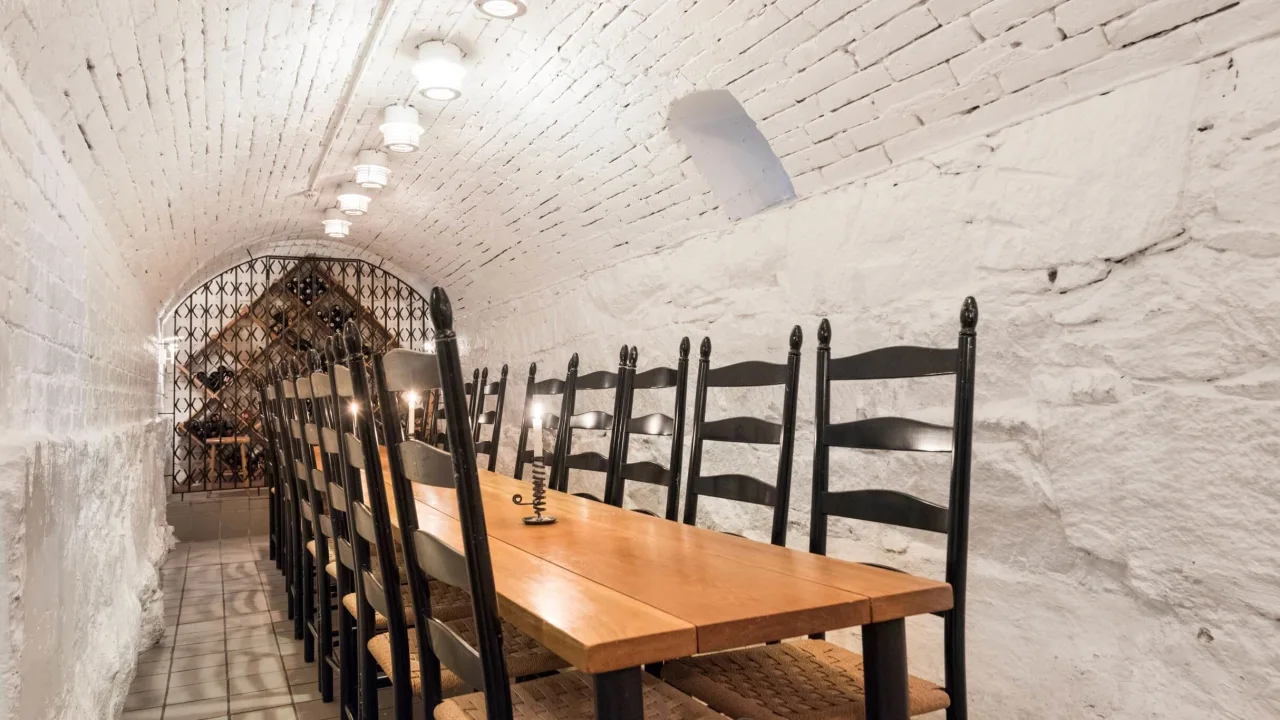 A long wooden table with black chairs lining each side, in a brick vaulted cellar with a wine rack and pendant lights.