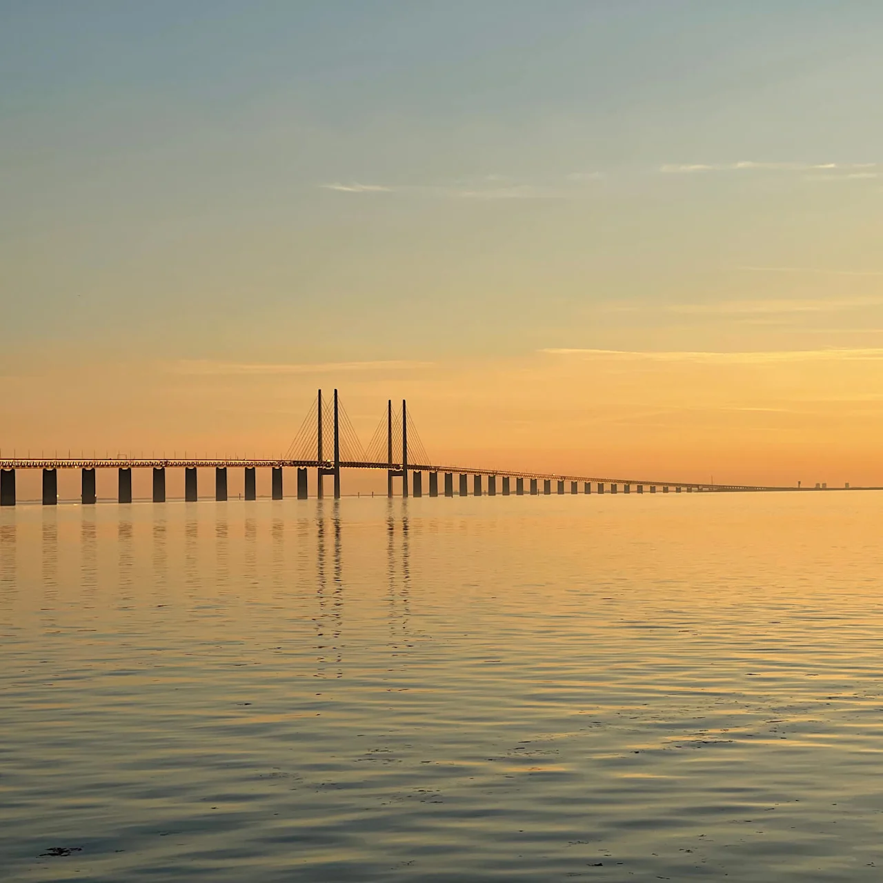 Broen strekker seg over rolig vann, reflekterer en solnedgangshimmel med oransje og blå toner, og skaper en fredelig atmosfære.