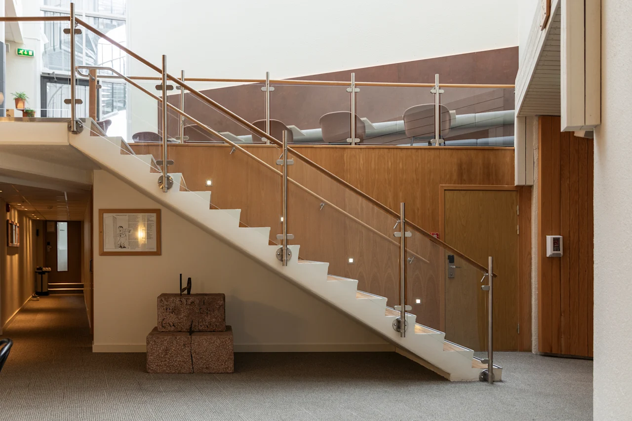 A modern interior with a white staircase featuring glass and wood railings, leading up to a seating area. A hallway is visible to the left.