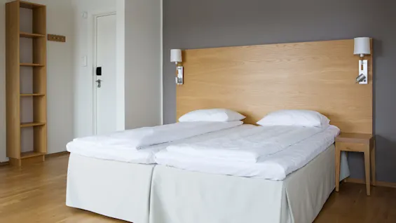 To pent, oppredde enkeltsenger ved siden av hverandre under et trehodegjerde, plassert i et minimalistisk rom med grå vegger, et lite trebord og en tom hylle.
