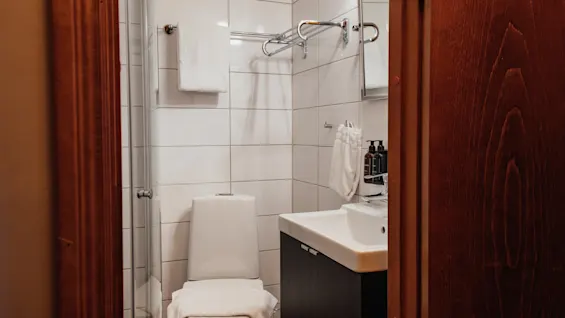 Ett ljust och prydligt hotellbadrum med vita kaklade väggar, en toalett, ett handfat med bekvämligheter och en glasinklädd dusch.