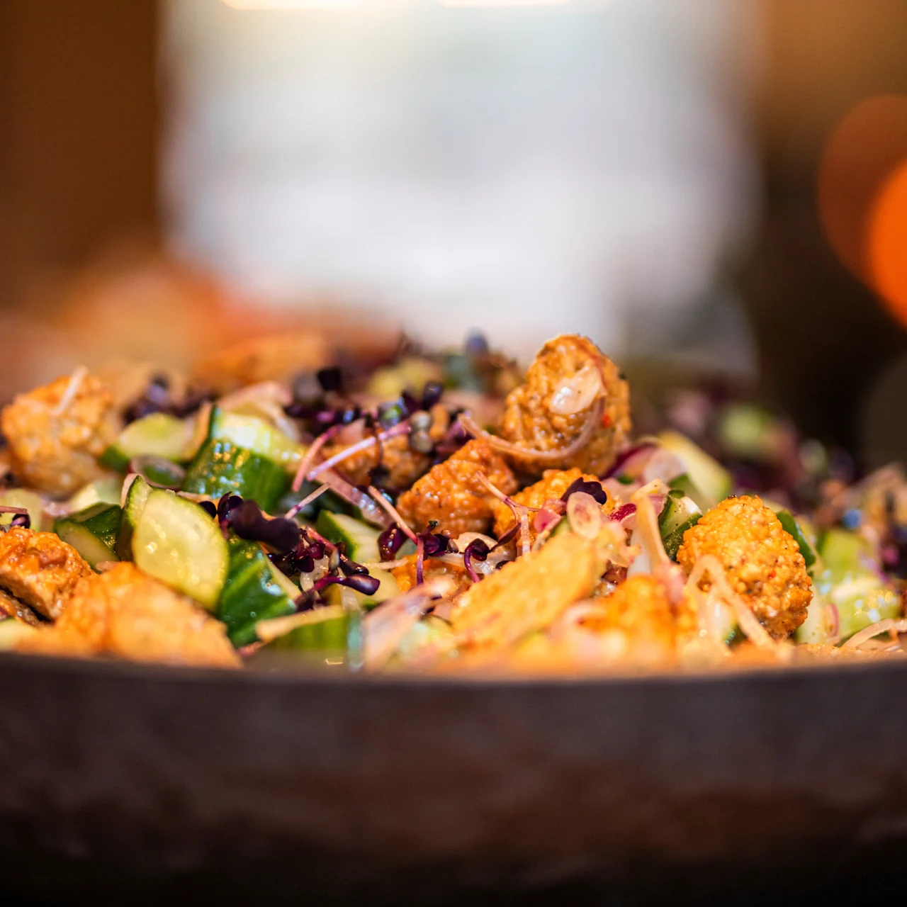 En farverig salat i en mørk skål indeholder blandede grøntsager, sprød tofu og drysset urter, placeret på et træbord med slørede varme lys i baggrunden.