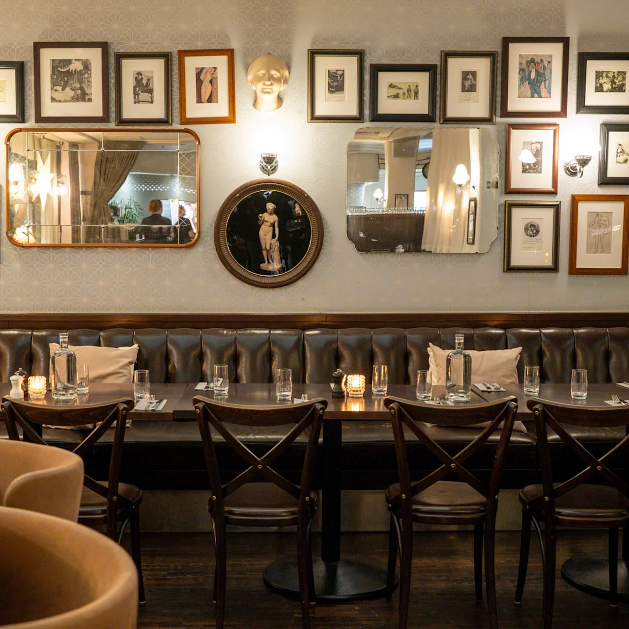 En svagt oplyst restaurantindretning har en lang, læderbetrukket bænk med pænt dækkede borde. Væggene viser mange indrammede kunstværker, spejle og dekorativ belysning.