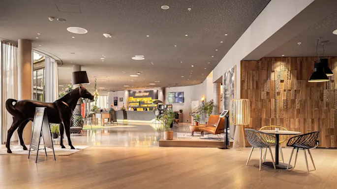 A horse statue with a lampshade head stands near a reception desk in an upscale lobby with modern seating and wooden decor. Sign reads "RECEPTION."