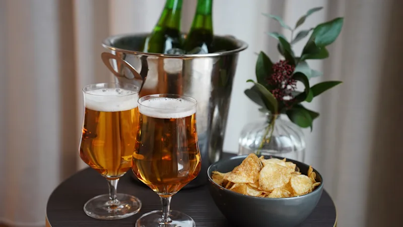 To ølglas fyldt med gylden væske og en skål med chips på et lille bord, ølflasker i en isspand og en plante i baggrunden.