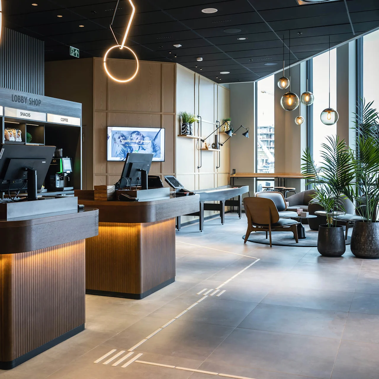 Reception borde står klar i en moderne lobby, oplyst af dekorative lys. Skiltet lyder "LOBBY SHOP: COLD DRINKS, SNACKS, COFFEE". Planter og siddepladser skaber en indbydende atmosfære.