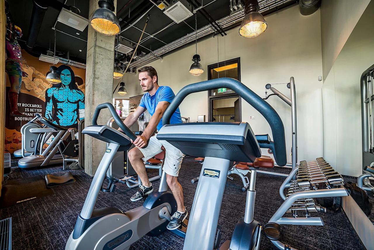 Man cycling on stationary bike in modern gym, surrounded by weights and equipment. Colorful wall art reads, "MY ROCK & ROLL IS BETTER THAN YOUR BODYBUILDING."