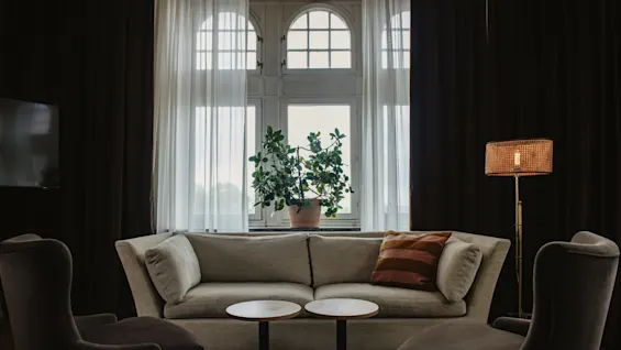 A beige sofa with cushions sits between two brown armchairs in a living room. A potted plant is on the windowsill, near sheer curtains and a wicker floor lamp.