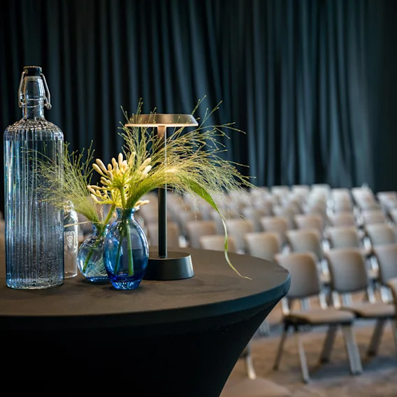 A table with a water bottle and floral decoration in a conference room with rows of empty chairs and a dark curtain.
