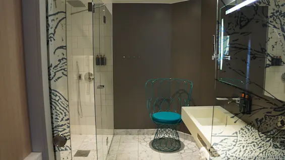 A modern bathroom with a glass-walled shower, marble floor, and a blue wire-frame chair.
