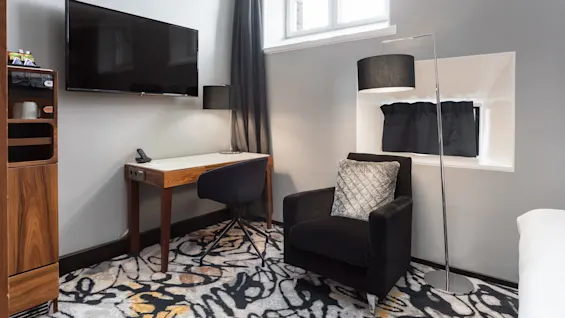 A modern styled hotel room with a mounted flat-screen TV, a desk with chair, armchair, floor lamp, and patterned carpet. 