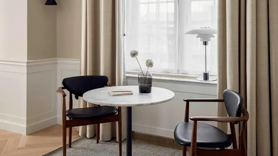 A round white marble table with wooden chairs sits by a window with beige curtains, decorated with a vase of flowers and a lamp, in a modern room.