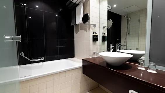 A modern bathroom features a white sink on a dark wooden counter, reflecting in a large mirror. Nearby, a bathtub with towels hanging above. The tiled walls enhance the sleek design.