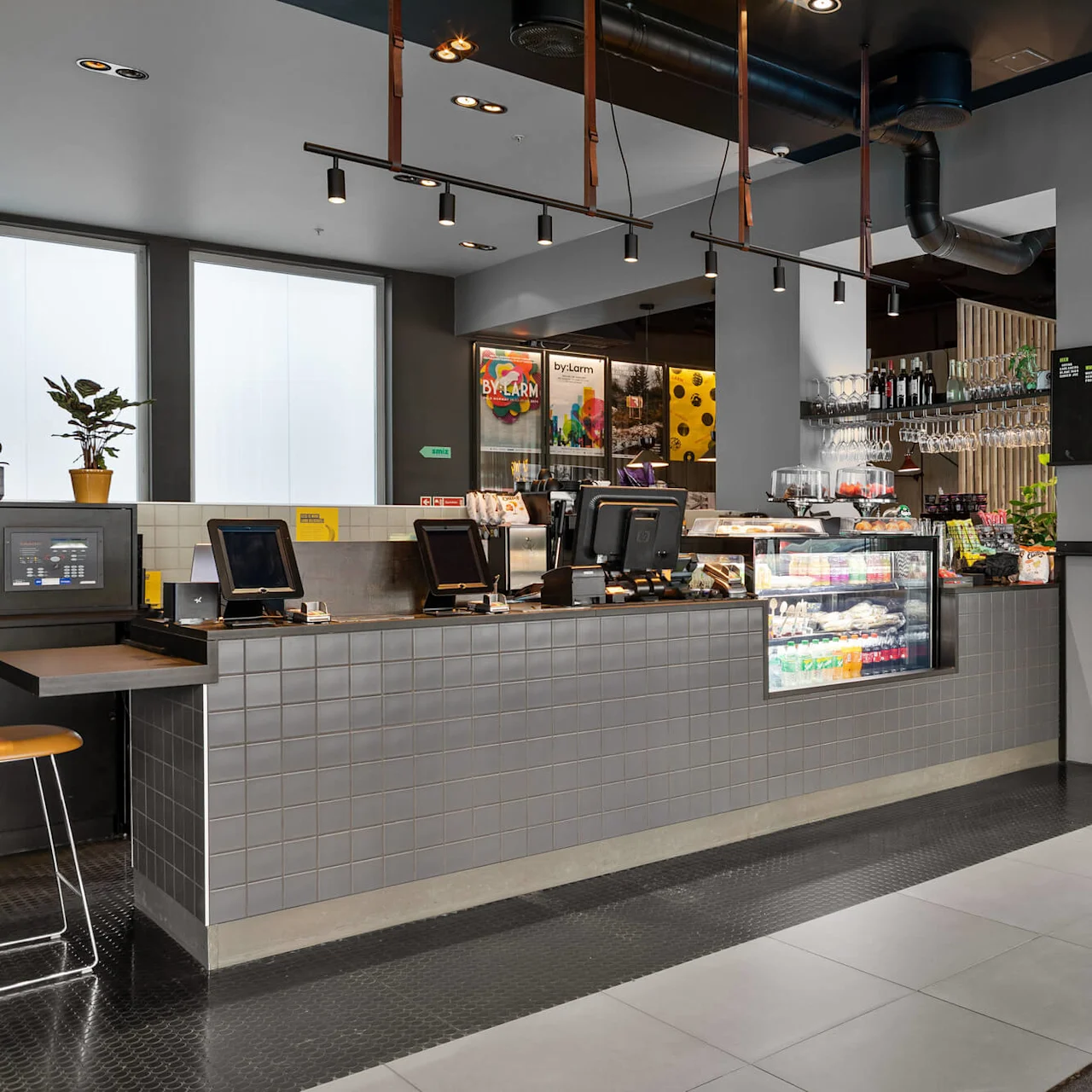 Café counter displaying food and beverages under glass, with digital registers; surrounded by modern decor and light fixtures. Visible signage: “BY:LARM.” Exit sign above door.