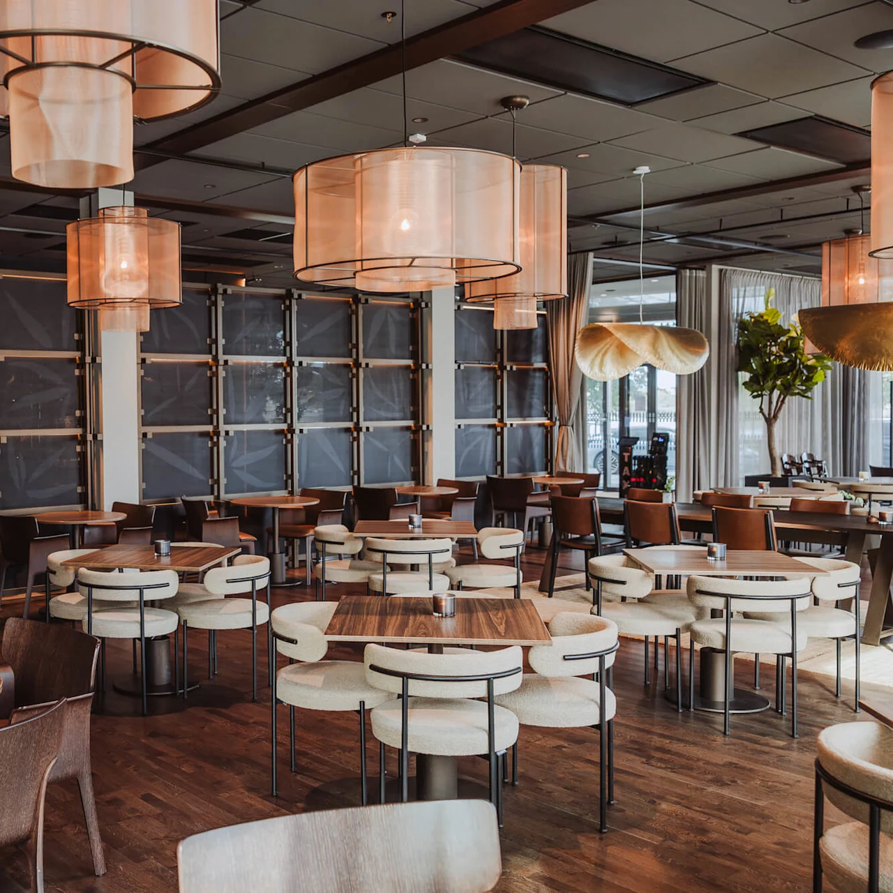 Rymlig restaurang med runda och fyrkantiga träbord omgivna av stolar under stora, moderna pendellampor. Träd och växter ger en naturlig atmosfär mot en stilren, samtida bakgrund.