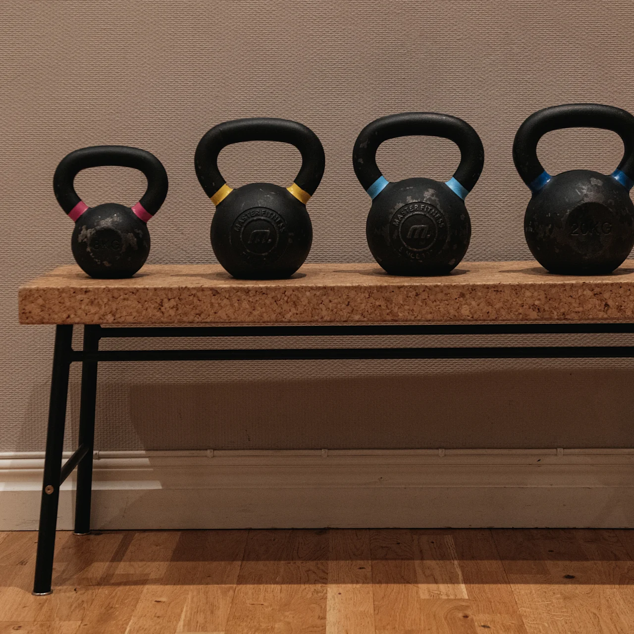 Kettlebells vilar på en träbänk, placerade mot en ljusbrun vägg på ett trägolv; varje kettlebell har ett färgat band på sitt handtag.