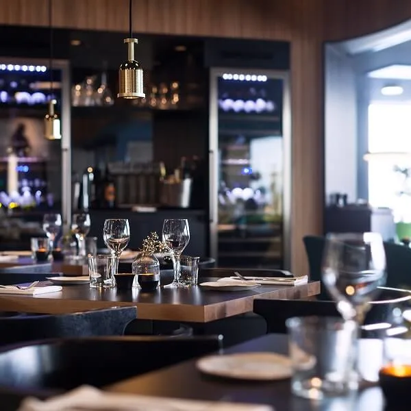 Borde dækket med glas og servietter i en hyggelig, blødt belyst restaurant, omgivet af trævægge og vinudstillinger, hvilket skaber en intim spiseatmosfære.