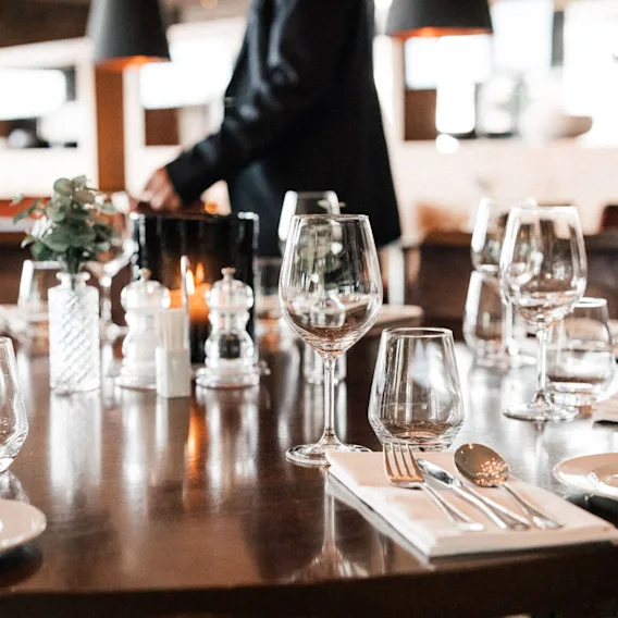 Elegant dekket restaurantbord med vinglass, bestikk, levende lys og en uskarp servitør i bakgrunnen.