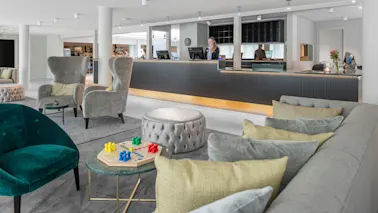 En moderne hotellobby med bløde stole og et lille bord, der rummer et brætspil. En receptionist står ved den glatte, sorte skranke. Lys, luftig indretning forstærker den rummelige atmosfære.