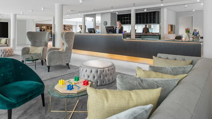 En moderne hotellobby har komfortable stoler og et lite bord med et brettspill. En resepsjonist står ved den stilige, svarte skranken. Lys, luftig innredning forbedrer den romslige atmosfæren.