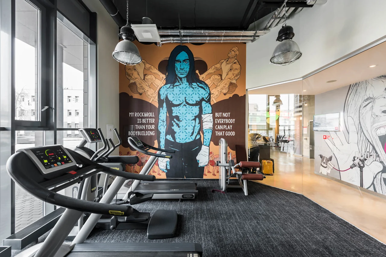 Treadmills sit stationary in a gym, facing a mural of a blue-skinned figure. Text reads: "MY ROCK&ROLL IS BETTER THAN YOUR BODYBUILDING BUT NOT EVERYBODY CAN PLAY THAT GOOD."