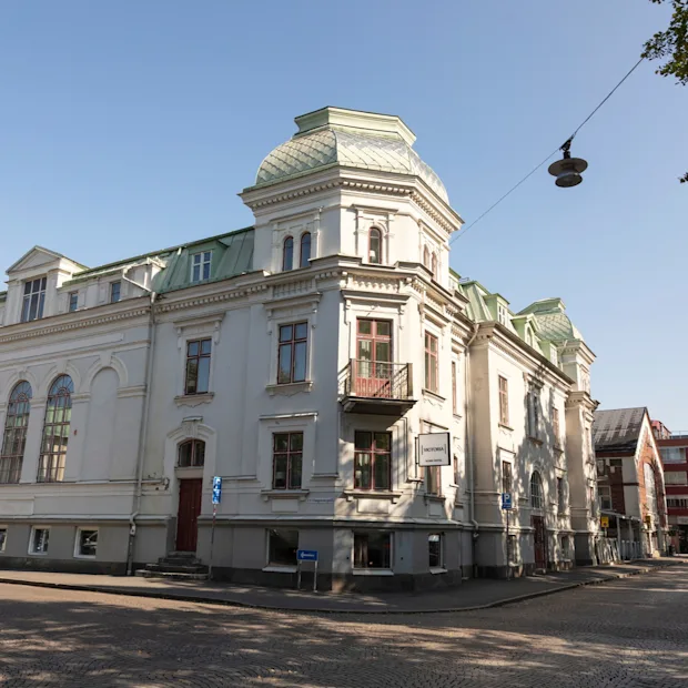 Upea rakennus, Home Hotel Victoria, jossa on erottuva katto ja arkkitehtonisia yksityiskohtia, seisoo mukulakivikadulla kirkkaan sinisen taivaan alla.