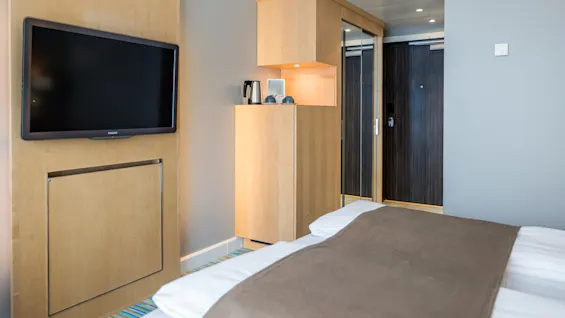 A wall-mounted Philips television faces a neatly made bed in a modern hotel room. A wooden cabinet beside it holds a kettle, cups, and a lit shelf, with a dark wooden door in the background.