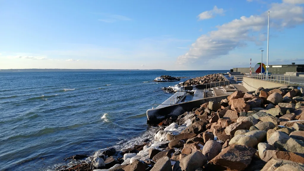En steinete kystlinje med en betongbrygge som strekker seg ut i det blå havet under en delvis skyet himmel. Litt is er synlig på steinene.